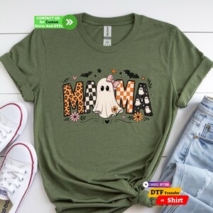 May include: An olive green t-shirt featuring a graphic print that spells MAMA. The letters incorporate leopard print, black and white checkers, and a ghost wearing a pink bow. The design includes small bats, stars, hearts, and flowers.