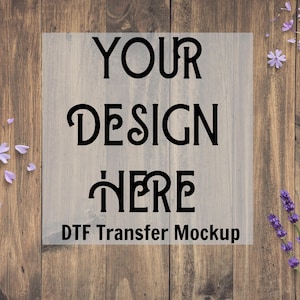 May include: A DTF Transfer Mockup with the words "YOUR DESIGN HERE" in a bold, black font. The text is on a light gray background, set against a rustic wooden backdrop. Lavender and purple flowers are scattered around the edges.