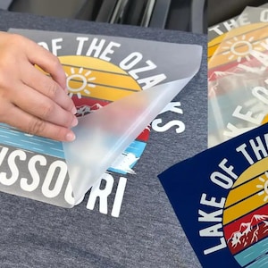 May include: A dark gray t-shirt featuring a screen-printed graphic of a sunset over mountains and water with the text Lake of the Ozarks Missouri. A clear transfer film is being peeled away from the design to reveal the colorful printed graphic.