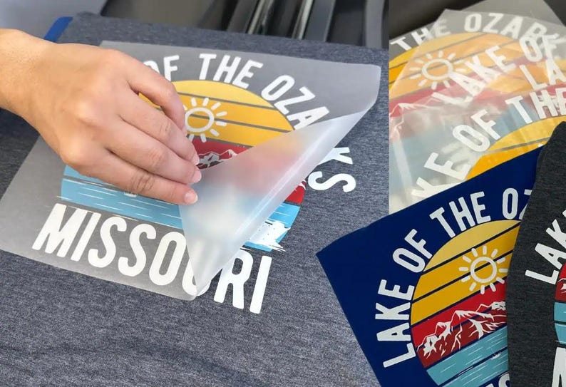 May include: A dark gray t-shirt featuring a screen-printed graphic of a sunset over mountains and water with the text Lake of the Ozarks Missouri. A clear transfer film is being peeled away from the design to reveal the colorful printed graphic.