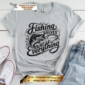 May include: A light gray short-sleeve t-shirt featuring a black graphic print of a fish on a hook with the text Fishing SOLVES everything. The shirt has a crew neckline and a standard fit, showcasing a detailed illustration of a bass fish.