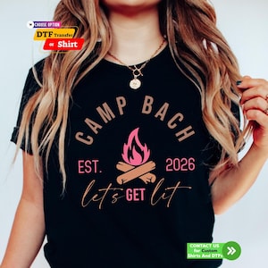 May include: A black t-shirt featuring a graphic design with the text "CAMP BACH" in tan, "EST. 2026" in pink, and "let's GET lit" in pink and tan. The center graphic depicts a stylized pink campfire with tan logs underneath the flames.
