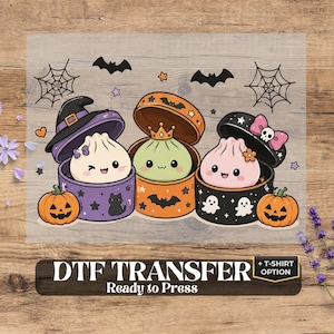 May include: A DTF transfer sheet featuring three cute cartoon dumplings in round Halloween-themed boxes. The dumplings are white, green, and pink, wearing a witch hat, crown, and bow. Includes pumpkins, bats, spiderwebs, and the text DTF TRANSFER Ready to Press + T-SHIRT OPTION.