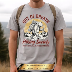 May include: A light gray t-shirt featuring a graphic of a bear wearing a backpack and holding a walking stick. The text reads OUT OF BREATH HIKING SOCIETY and DON'T WORRY, I'LL BE THERE IN A MINUTE. A banner at the bottom reads DTF TRANSFER or PRINTED T-SHIRT.