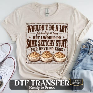May include: A beige short-sleeved t-shirt featuring a brown graphic print that reads: WOULDN'T DO A LOT for turkey or ham BUT I WOULD DO SOME SKETCHY STUFF FOR DEVILED EGGS, accompanied by an illustration of three deviled eggs.