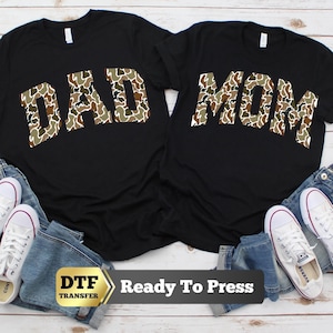May include: Two black t-shirts featuring the words DAD and MOM in a camouflage print. The shirts are displayed with blue denim jeans and white canvas sneakers. A gold-toned label at the bottom displays the text DTF TRANSFER Ready To Press.