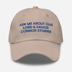 May include: A beige baseball cap with a curved brim. The cap features the embroidered text "ASK ME ABOUT OUR LORD & SAVIOR CONNOR STORRIE" in blue, centered on the front. The cap has a structured crown and an adjustable strap.