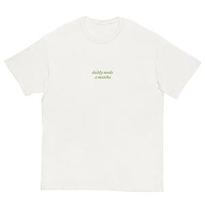 May include: White t-shirt with the text "daddy needs a matcha" in green script. The shirt has short sleeves and a crew neck. The text is centered on the chest.
