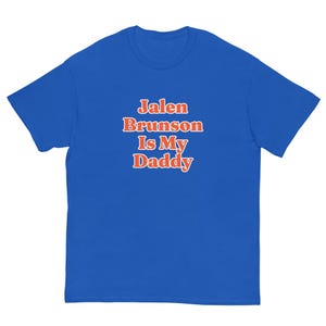 Op de afbeelding: Een koningsblauw T-shirt met korte mouwen en de tekst Jalen Brunson Is My Daddy in oranje letters met een witte omlijning, gecentreerd op de borst. Het kledingstuk heeft een ronde hals en een standaard pasvorm, gemaakt van effen stof.