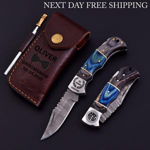 May include: A set of two folding knives with blue and gray handles, a leather sheath, and a sharpening rod. The knives have a patterned blade and are engraved with "25 YEARS TOGETHER" and initials. The sheath is engraved with "OLIVER" and "GROOMSMAN 08.04.2022".