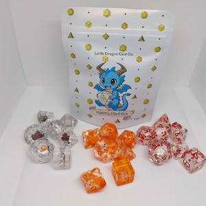 May include: A white bag with a cartoon dragon illustration and the text "Little Dragon Dice Co." and "Mystery Filled Dice." Assorted polyhedral dice in clear, orange, and red colors are scattered in front of the bag.