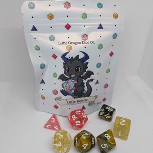 Op de afbeelding: Een witte zak met een cartoon draak die een veelkleurige dobbelsteen vasthoudt, met de tekst "Little Dragon Dice Co." en "Little Rejects". Verschillende veelzijdige dobbelstenen in verschillende kleuren liggen verspreid voor de zak.