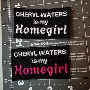 May include: Two rectangular black embroidered patches with white borders. The top patch features white text reading CHERYL WATERS is my Homegirl. The bottom patch features white text reading CHERYL WATERS is my, with Homegirl in pink thread.