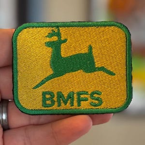 May include: Embroidered patch featuring a green deer leaping across a yellow background. The patch has a green border and the letters "BMFS" are at the bottom. The patch is rectangular with rounded corners.