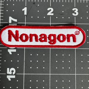 May include: An embroidered patch with a white background and a red border, featuring the red text "Nonagon" followed by a registered trademark symbol. The patch is positioned on a black cutting mat with white grid lines measuring 7.62 cm wide.