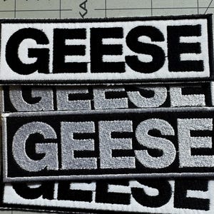 May include: Embroidered patches with the word "GEESE" in bold, black capital letters on a white background. The patches are rectangular and have a black border. The text is also available in silver.
