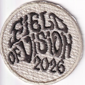 May include: A circular embroidered patch with a white background and a dark gray stitched border. The patch features the dark gray embroidered text "FIELD VISION 2026" in a stylized, wavy font centered on the white fabric surface.