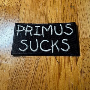 May include: Black rectangular patch with the words "PRIMUS SUCKS" embroidered in white. The patch is likely designed to be sewn or ironed onto clothing or accessories. The text is in a bold, sans-serif font.