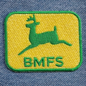 May include: A square embroidered patch with rounded corners featuring a green leaping deer silhouette centered on a yellow background. The text BMFS is embroidered in green capital letters below the deer. The patch has a green border.