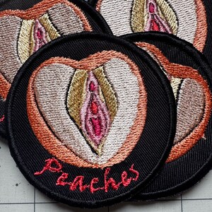 Puede incluir: Parches bordados con fondo negro y diseño en forma de melocotón. El diseño presenta una forma de vulva rosa y beige con detalles en naranja y beige. La palabra "Peaches" está bordada en escritura roja.