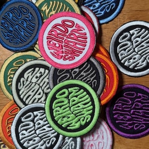 May include: A collection of embroidered patches with the words "WEIRDO SWARM" in a circular design. The patches feature various color combinations, including blue, pink, green, and purple, with white or black lettering. The patches are round and have a stitched border.