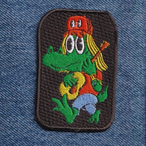 May include: A rectangular embroidered patch featuring a green cartoon alligator with long blonde hair wearing a red baseball cap, a red shirt, and blue shorts. The alligator is holding a yellow acoustic guitar and has a guitar neck on its back.