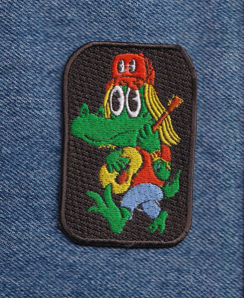 Op de afbeelding: Een rechthoekige geborduurde patch met een groene cartoon-alligator met lang blond haar, een rode honkbalpet, een rood shirt en een blauwe broek. De alligator houdt een gele akoestische gitaar vast en draagt een gitaarhals op de rug.
