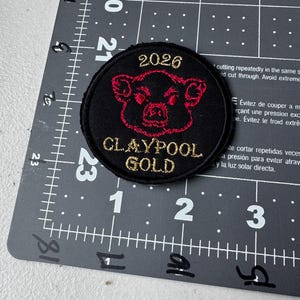 May include: A round black patch with a red pig face design and gold text that reads "2026" and "CLAYPOOL GOLD". The patch is on a gray cutting mat with measurement markings.