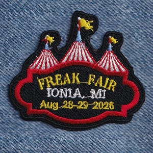 May include: An embroidered patch featuring three red and white striped circus tents with yellow flags. The black center has red trim and yellow text reading FREAK FAIR, IONIA, MI, and Aug 28-29-2026 in white and yellow embroidery thread.