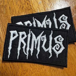 Peut inclure: Deux écussons rectangulaires noirs avec le mot "PRIMUS" brodé en blanc, en lettres stylisées. Les écussons sont posés sur une surface en bois. Les écussons sont probablement destinés aux vêtements ou aux accessoires.