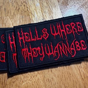 May include: Three black rectangular patches with red embroidered text that reads "Hells Where They Wanna Be". The patches are stacked on a wooden surface.