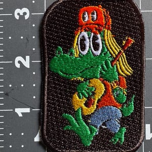 Op de afbeelding: Een rechthoekige geborduurde patch op een donkerbruine achtergrond met een groene alligator die een oranje pet draagt en op een gele gitaar speelt. De patch ligt naast een liniaal met een hoogte van ongeveer 7,6 cm.