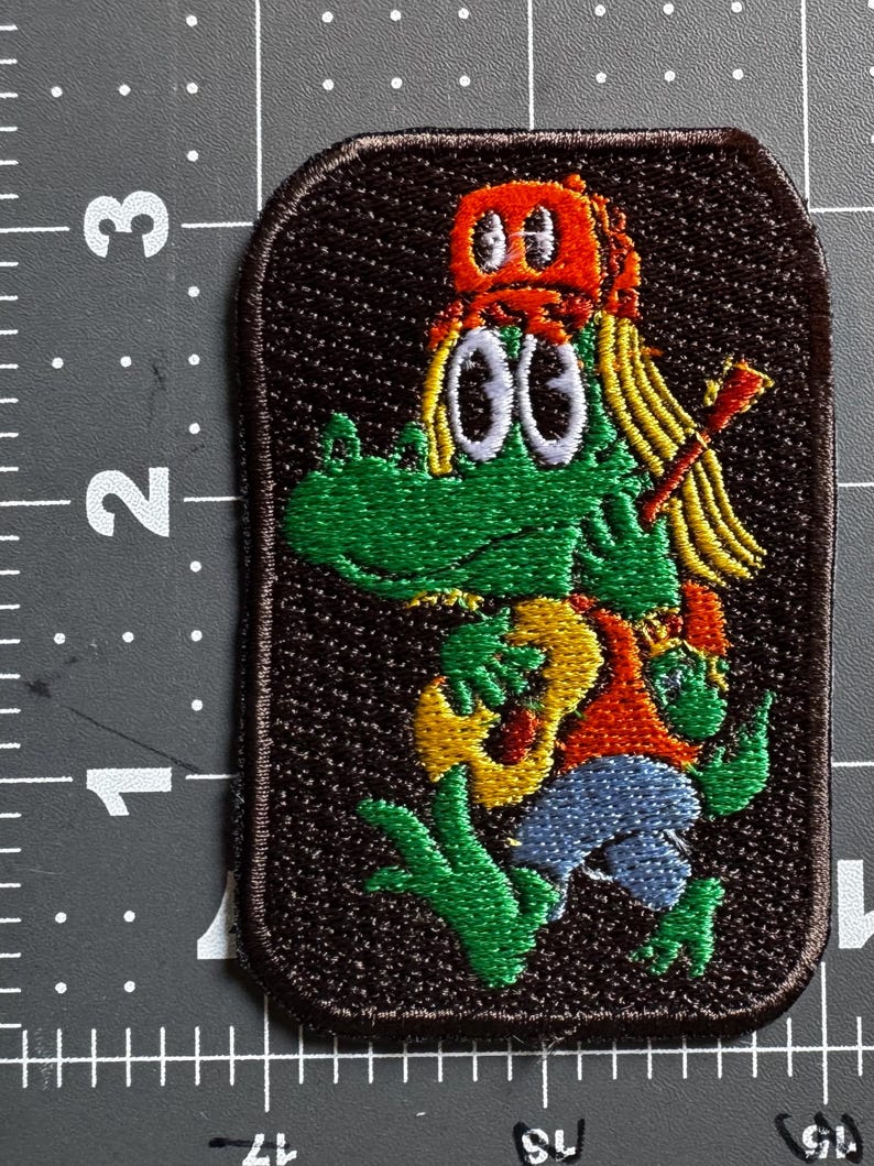 Op de afbeelding: Een rechthoekige geborduurde patch op een donkerbruine achtergrond met een groene alligator die een oranje pet draagt en op een gele gitaar speelt. De patch ligt naast een liniaal met een hoogte van ongeveer 7,6 cm.
