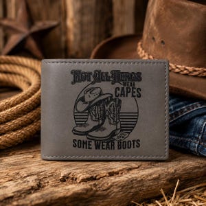 May include: Gray leather wallet with the text "Not All Heroes Wear Capes, Some Wear Boots." The wallet features an illustration of a cowboy hat and boots. The background includes a cowboy hat and rope.