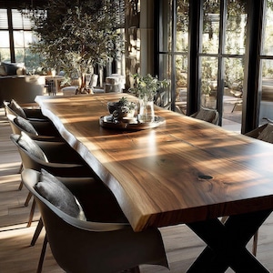 May include: A substantial wooden dining table with a natural edge and a warm brown hue. The table, supported by a black metal base, is surrounded by modern chairs with grey cushions. A decorative tray with plants and glassware sits on the table.
