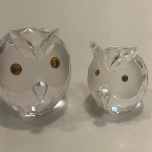 May include: Two clear crystal owl figurines. The owls have faceted bodies, with small, round, amber-colored eyes. One owl is larger than the other. The owls are decorative items.