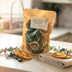 May include: A clear cellophane bag filled with dried orange slices, green leaves, and other botanicals. The bag has a label that reads "Rectful Moments." Several dried orange slices are arranged on a white surface with a folded linen cloth.