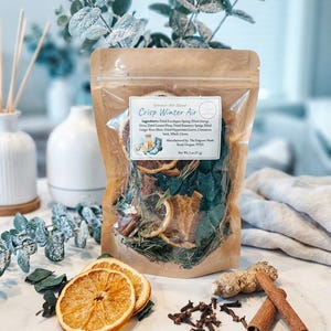 May include: A clear bag of potpourri blend labeled "Crisp Winter Air" with dried orange slices, cinnamon sticks, and cloves. The ingredients include dried eucalyptus, rosemary, and ginger root. The bag is displayed with eucalyptus branches and a white vase.