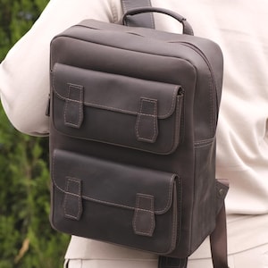 May include: A dark brown rectangular backpack featuring two stacked front pockets with flap closures, a top handle, and adjustable shoulder straps. The bag has a smooth, matte finish and is worn on the back with a single strap visible over the shoulder.