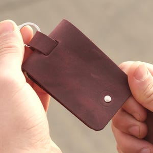 Puede incluir: Un llavero rectangular de cuero marrón oscuro con acabado mate liso, una anilla de metal plateado sujeta a una trabilla de cuero en la parte superior y un pequeño remache de metal plateado centrado cerca del borde inferior del cuero.