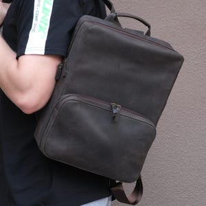 May include: A dark brown leather backpack with a rectangular shape, featuring a large main compartment, a smaller front zippered pocket, a top handle, and adjustable shoulder straps. The bag has a matte finish and dark metal zipper pulls.