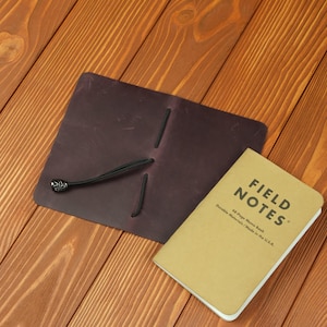 Puede incluir: Una funda de cuero marrón oscuro para cuaderno con bandas elásticas negras y una cuenta metálica decorativa, acompañada de un cuaderno Field Notes color canela con el texto Field Notes 48-Page Memo Book Durable Materials Made in the U.S.A. impreso.