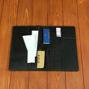 Pode incluir: Uma carteira de viagem de couro preto com vários compartimentos e bolsos internos. A carteira contém um passaporte azul, um caderno bege com a palavra FIELD impressa na capa, documentos de papel branco e um pequeno cartão branco com detalhes.
