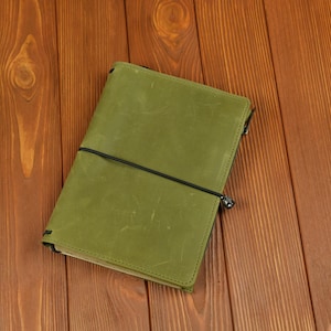 May include: A rectangular olive green leather notebook cover featuring a smooth, matte finish with visible surface scuffs and scratches. The cover is secured by a thin black elastic cord that wraps horizontally around the center of the notebook.