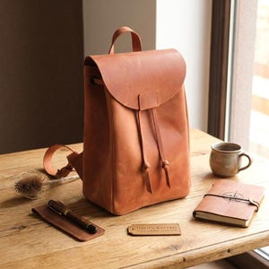 May include: A tan leather backpack with a fold-over flap, drawstring closure, and top handle. Beside it are a brown leather pen case with a pen, a small rectangular tag labeled HANDCRAFTED FULL GRAIN LEATHER, a notebook with a compass design, and a mug.