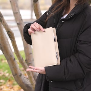 May include: A beige leather notebook with a pen in the spine. The notebook is held in front of a blurred outdoor background. The notebook is rectangular and appears to be closed.