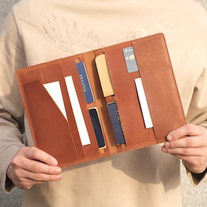 May include: A tan leather travel wallet featuring multiple vertical slots and pockets. The interior contains a blue passport with the text UKRAINA, a dark blue card holder, a beige card, a grey credit card, and several white paper inserts.