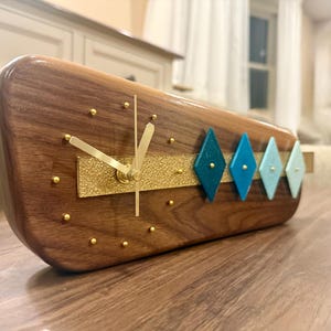 May include: A horizontal wooden clock with a polished dark brown finish, featuring gold-toned hands and small gold dot hour markers. A textured gold horizontal strip runs across the center, accented by four diamond-shaped ornaments in blue shades.