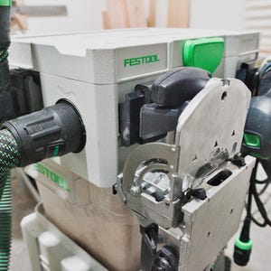 May include: A close-up of a gray and green power tool system. The system includes a gray box with the brand name "FESTOOL" in green, a green and black hose, and a silver metal component.