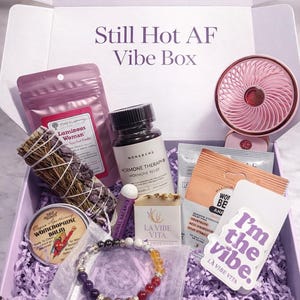 May include: A lavender-colored gift box labeled "Still Hot AF Vibe Box" filled with self-care items. Includes a pink fan, supplements, sage bundle, bracelet, soap, and sticker that says "I'm the vibe."